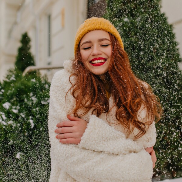 cheerful-ginger-lady-posing-under-snow-smiling-lovable-woman-in-knitted-hat-standing-near-spruce