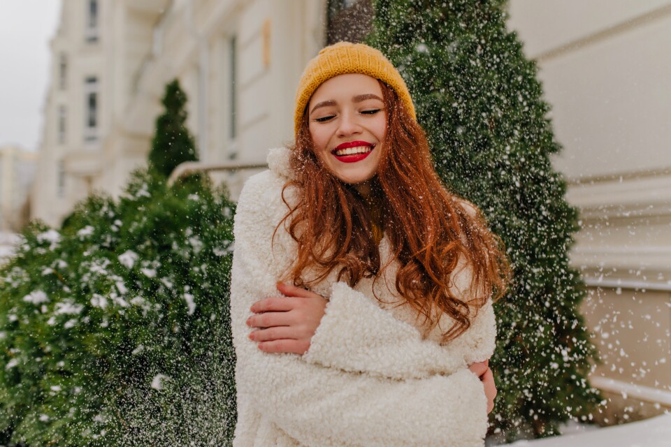 cheerful-ginger-lady-posing-under-snow-smiling-lovable-woman-in-knitted-hat-standing-near-spruce